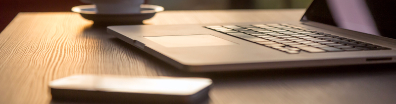 A laptop, coffee mug and phone sit on a desk and reflect the sunset.